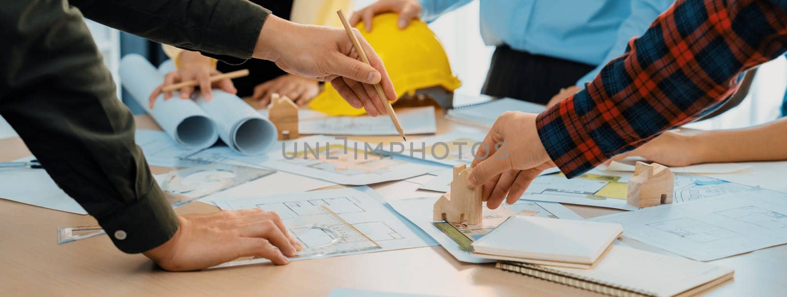 Professional architect team and safety engineer with safety helmet brainstorming about architectural project on meeting table with blueprint scatter around. Close up. Focus on hand. Delineation.