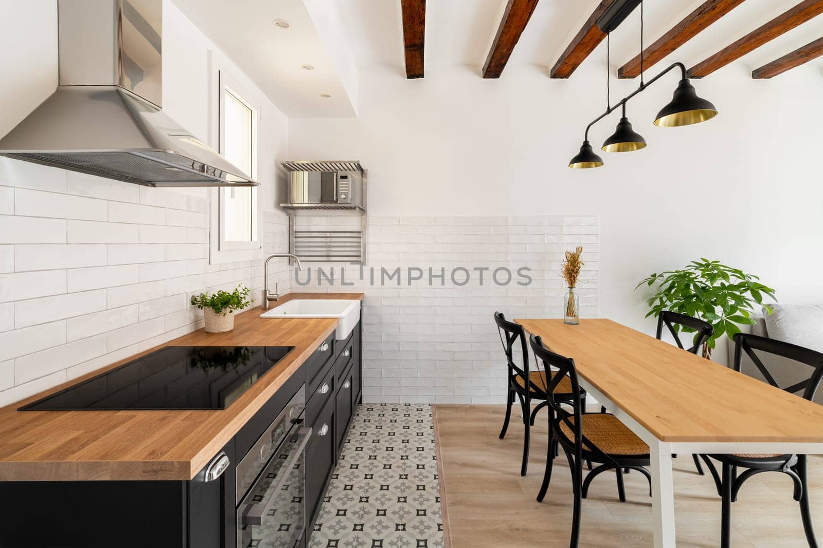 Long dining table with four chairs next to a kitchen island with a build in stove and extractor hood with white walls and renovation beams in a newly built house. Copyspace by apavlin