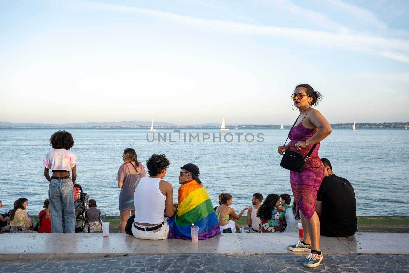 Lisbon, Portugal. 17 June 2023 Friends spending time together at Pride Parade. People communicating sitting on promenade with ocean and sailboats background. Rainbow flag for support LGBT individuals