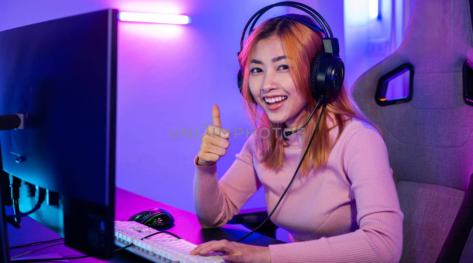 Gamer playing online game wear gaming headphones looking to camera expressing success with game giving thumbs up sign, Smiling woman live stream she play video game at home neon lights living room