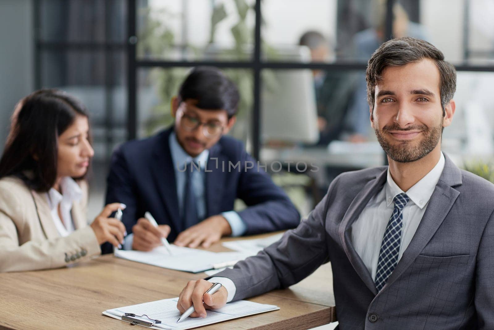 young man posing for the camera while sitting at a table in the office against the background of his colleagues