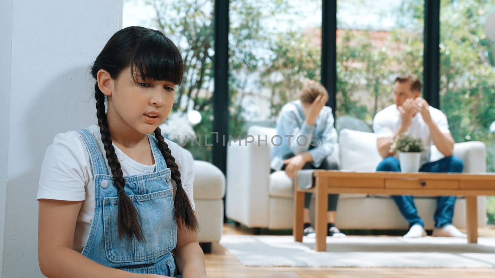 Stressed and unhappy young girl huddle in corner, cover her ears blocking sound of her parent arguing in background. Domestic violence at home and traumatic childhood develop to depression. Synchronos