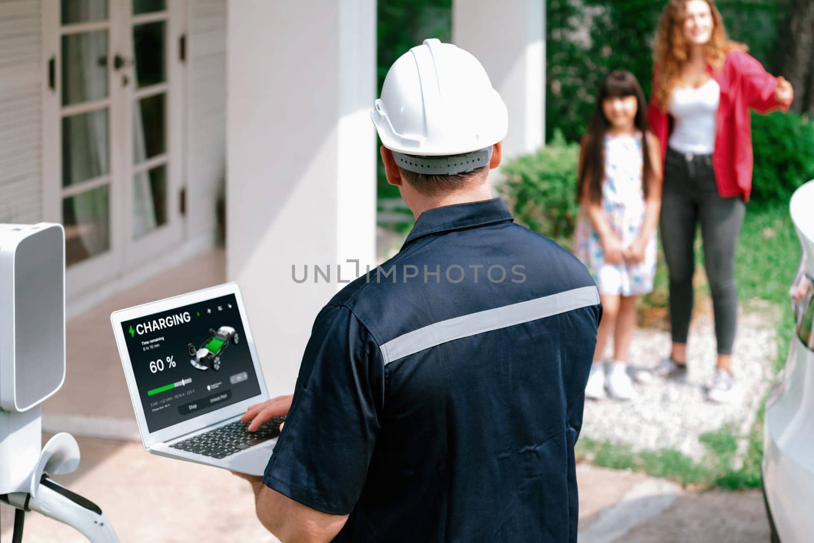 Qualified technician working on home EV charging station installation, making troubleshooting and configuration setup on charging system with laptop for EV at home with the family present. Synchronos