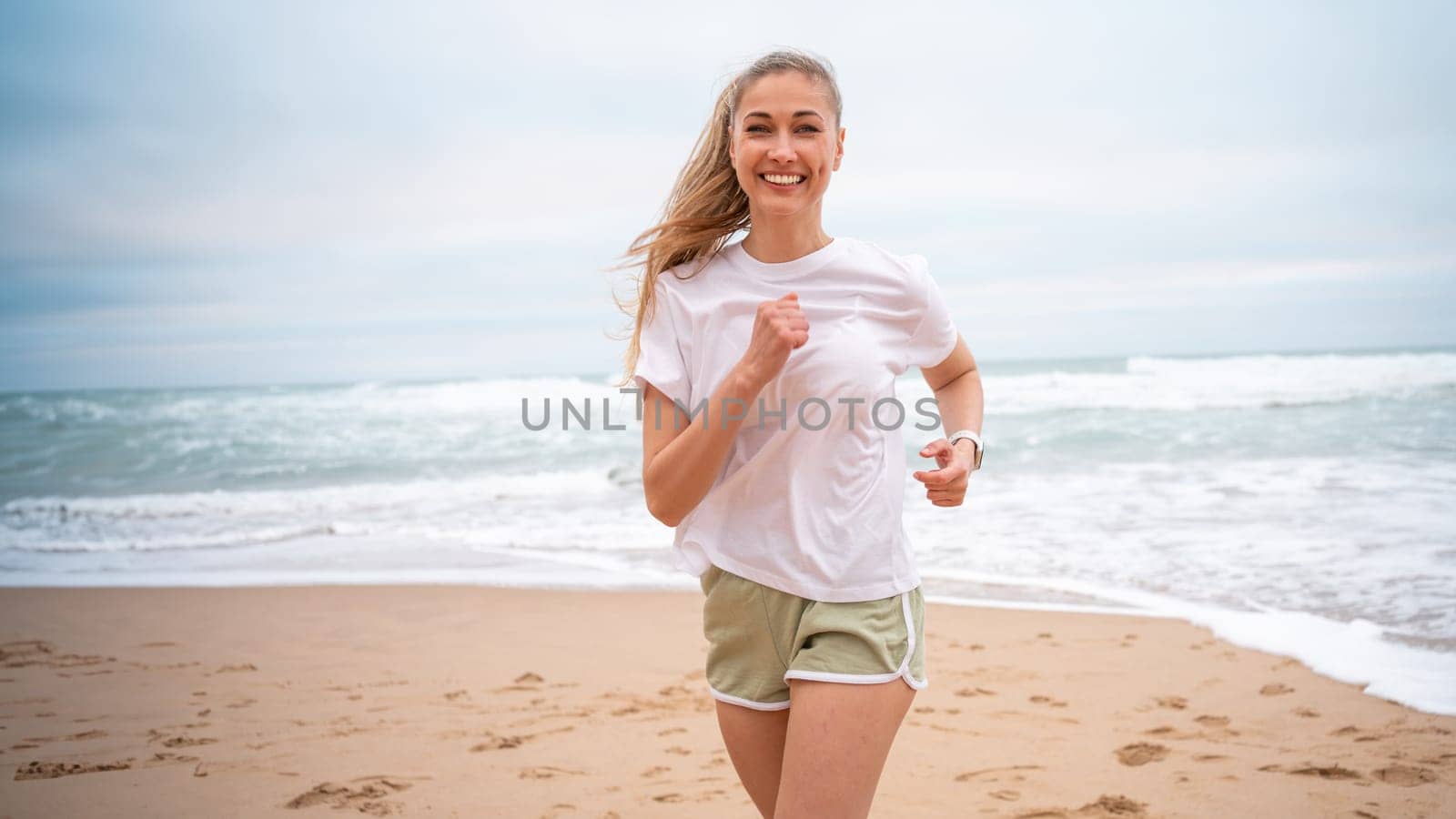 Smiling female athlete jogging on sandy sea beach. Fit woman in white top and sporty shorts. Active lady running along shoreline represents healthy lifestyle.