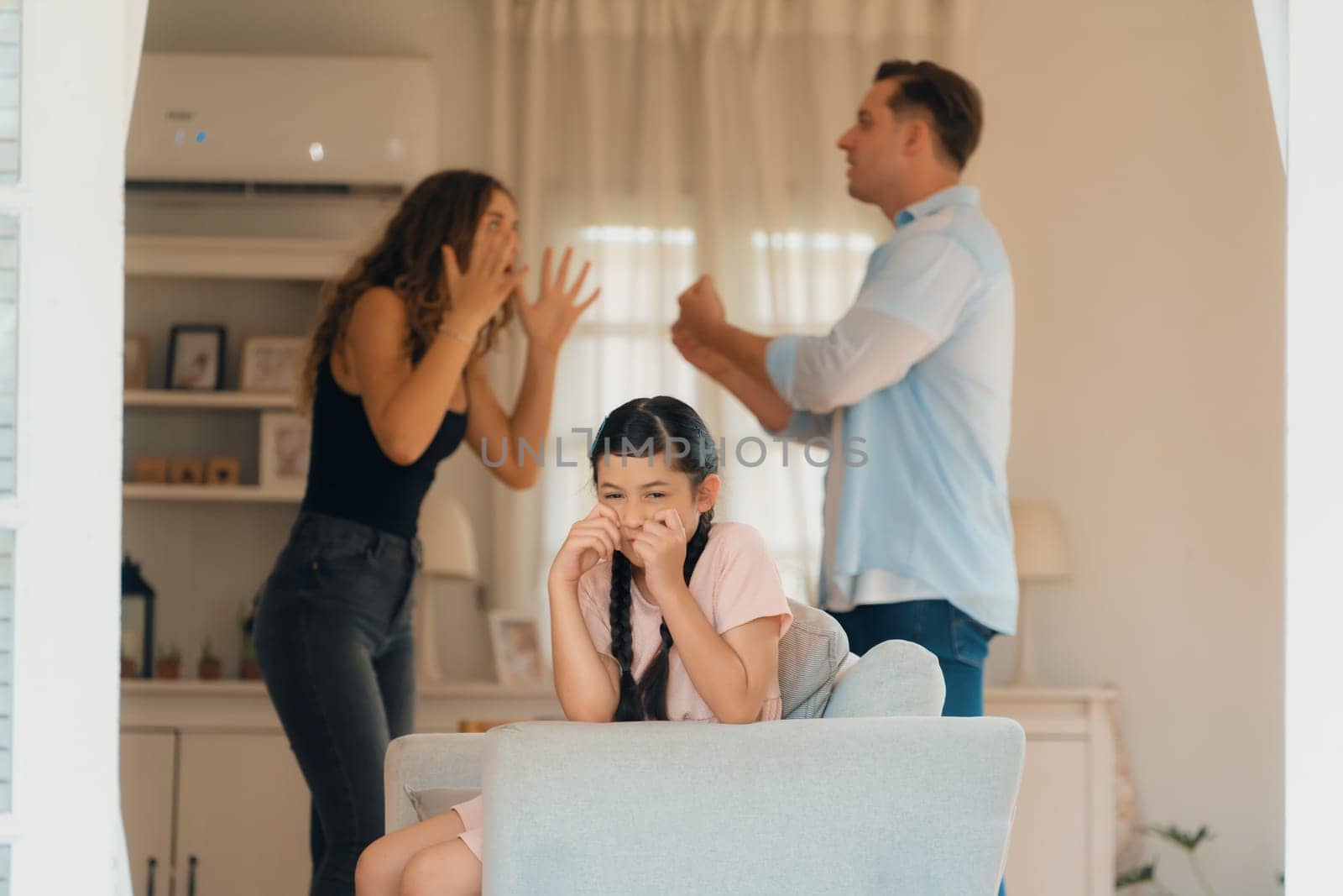 Annoyed and unhappy young girl sitting on sofa trapped in middle of tension by her parent argument in living room. Unhealthy domestic lifestyle and traumatic childhood develop to depression.Synchronos