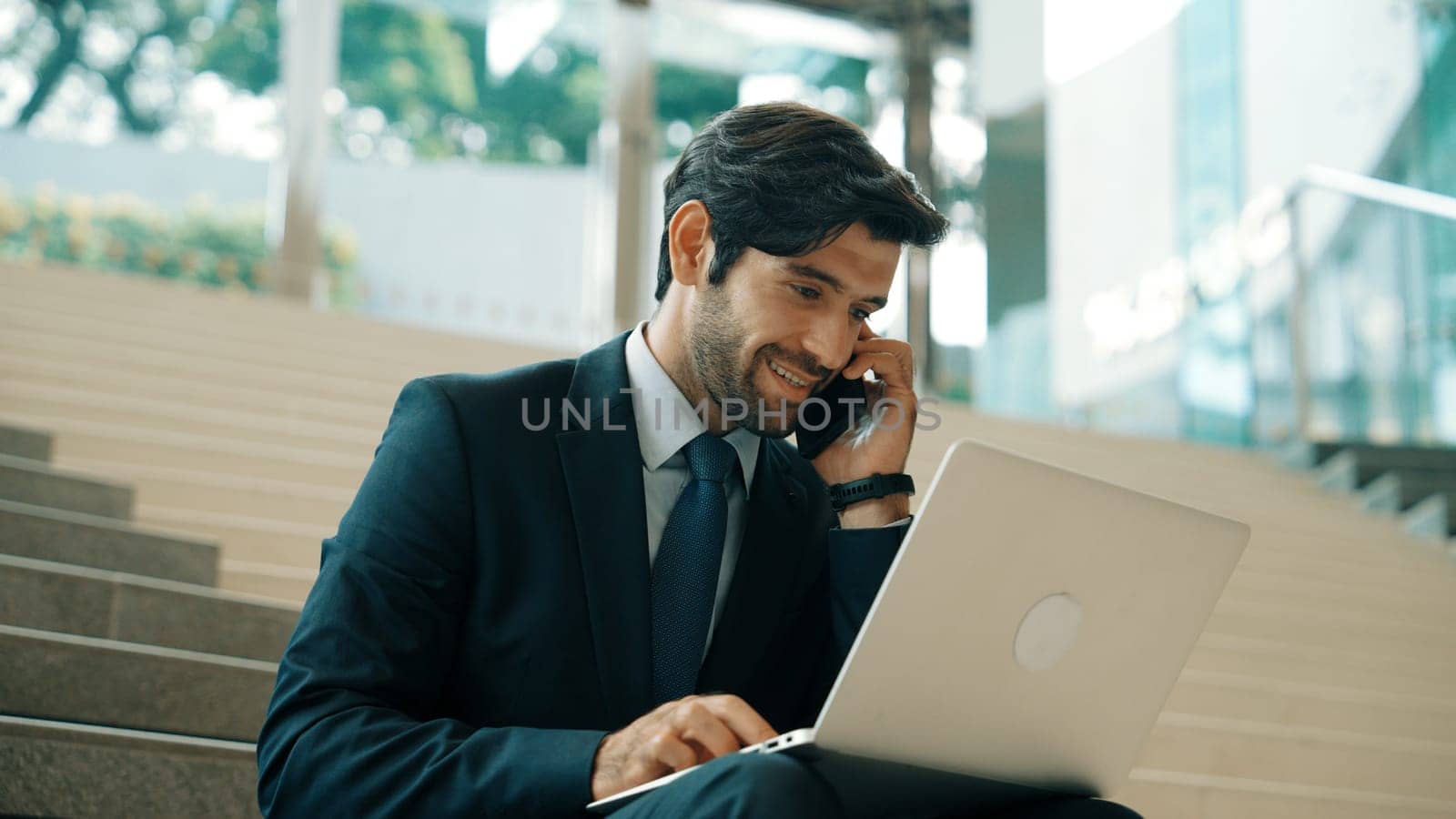 Skilled investor working or planing strategy by using laptop at stair. Professional business man wearing suit while working and typing data analysis by using laptop and talking to phone. Exultant.