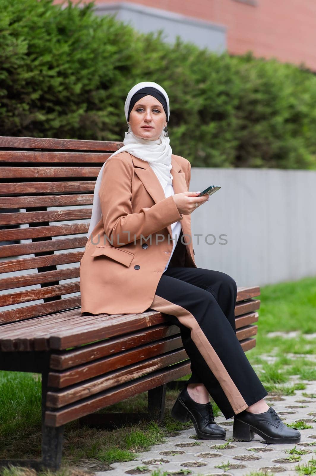 Young woman in hijab uses a smartphone while sitting on a bench outdoors. by mrwed54