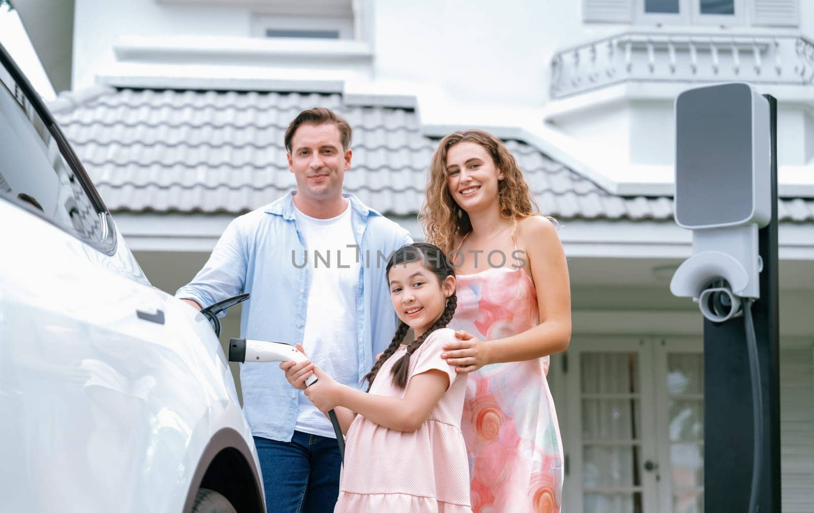 Happy little young girl learn about eco-friendly and energy sustainability as she help her family recharge electric vehicle from home EV charging station. EV car and modern family concept. Synchronos