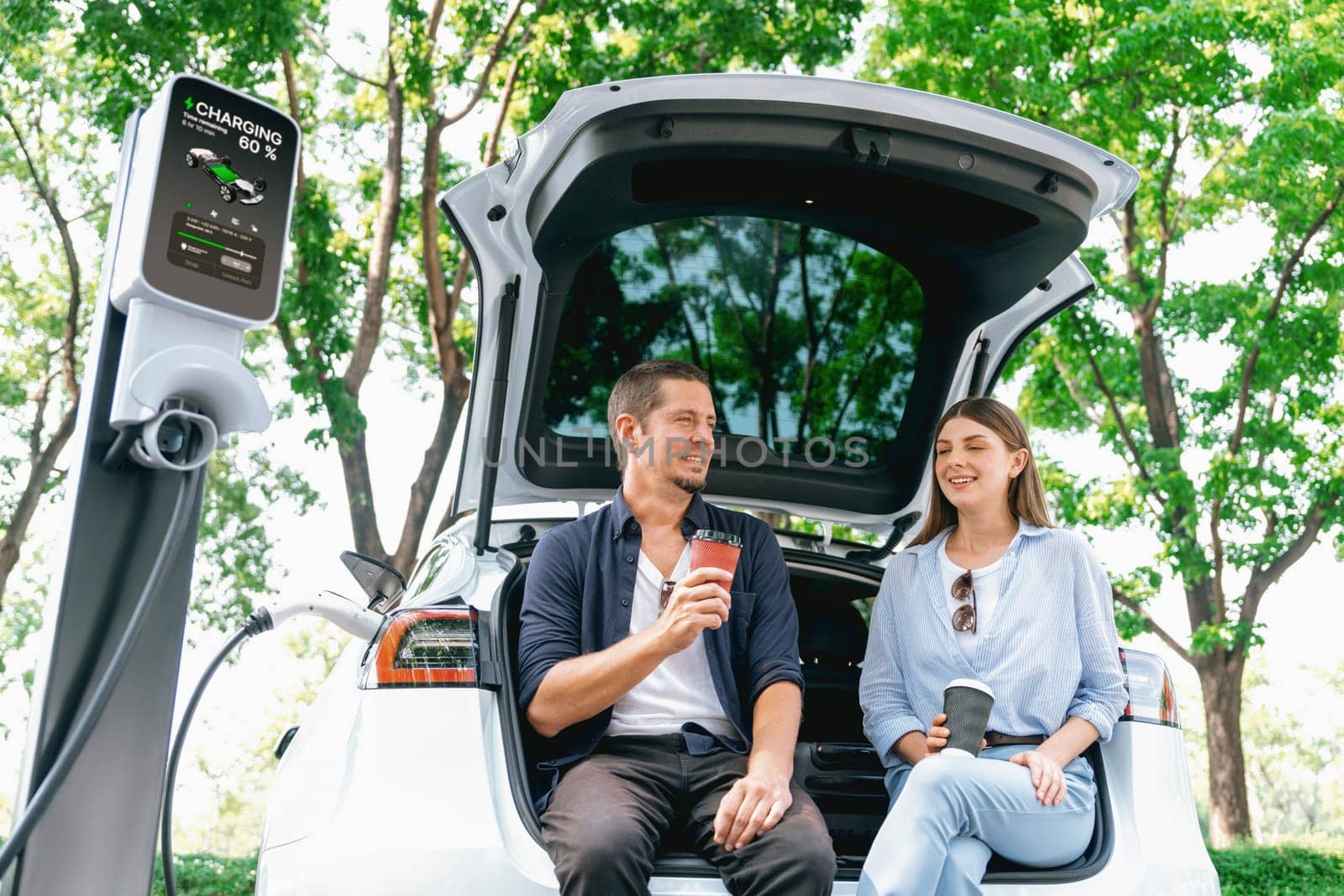 Lovely young couple drinking coffee while recharging battery for electric car during road trip travel EV car in natural forest or national park. Eco friendly travel during vacation and holiday. Exalt