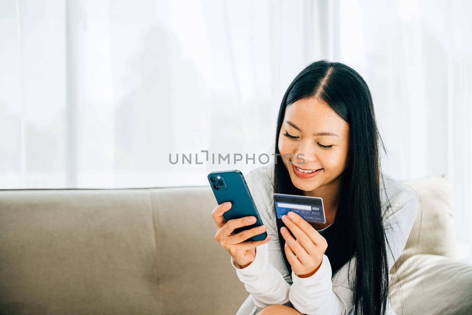 Smiling woman holds smartphone and credit card shops online on sofa by Sorapop