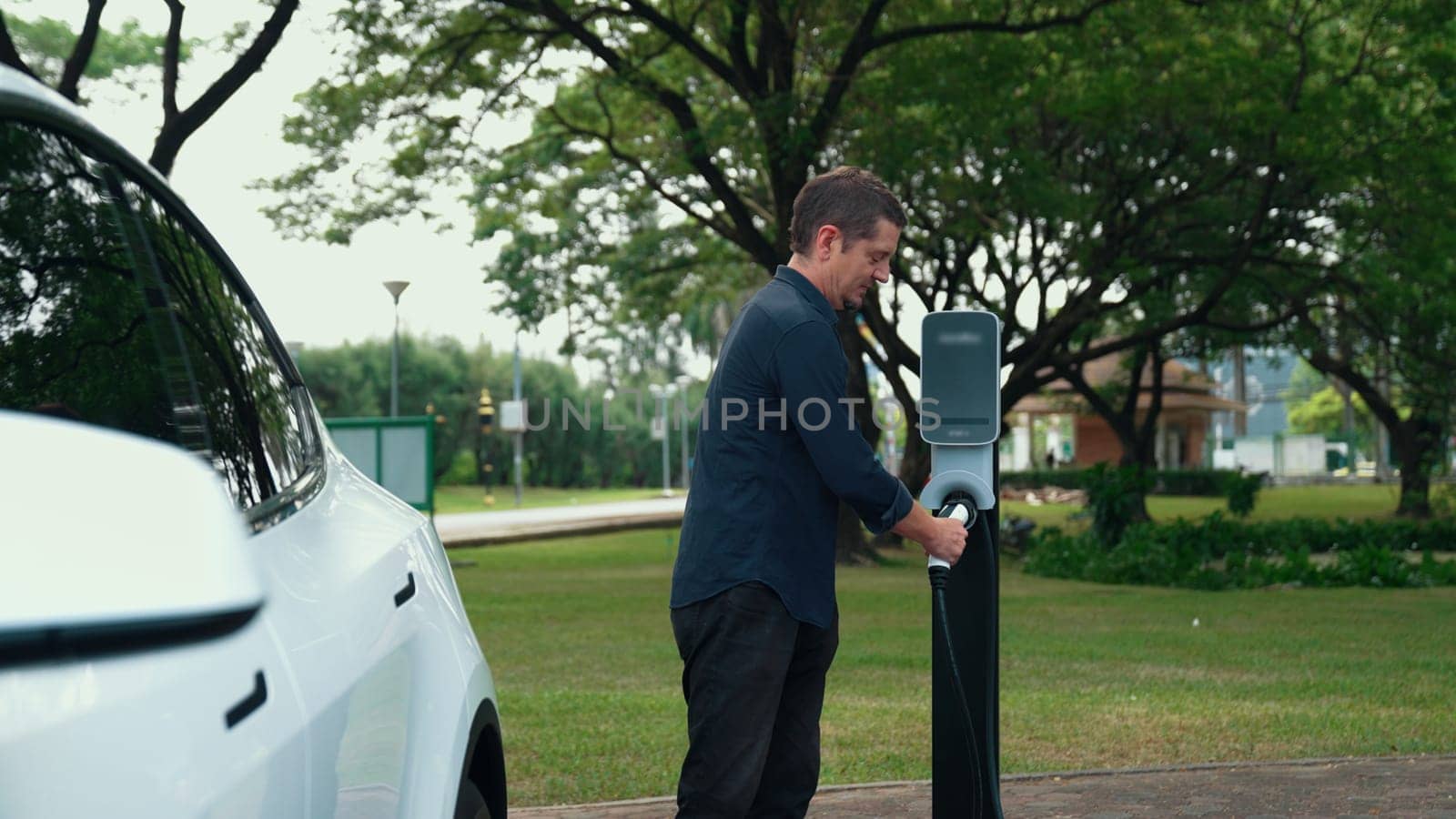 Man recharging battery for electric car during road trip travel EV car in natural forest or national park. Eco friendly travel during vacation and holiday. Exalt