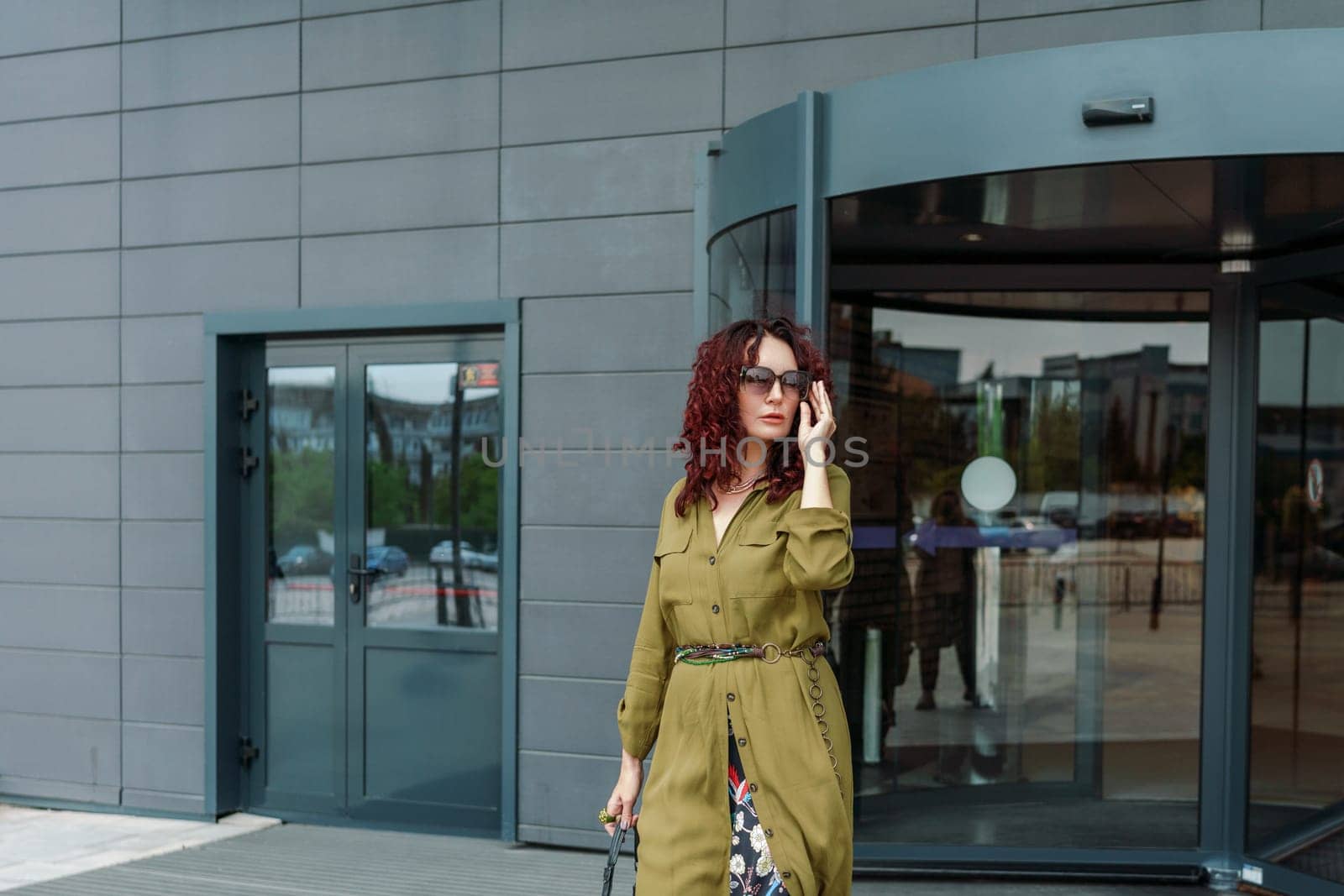 Woman leaves a supermarket. Caucasian model with long brunette hair, wears sunglasses and a khaki dress.