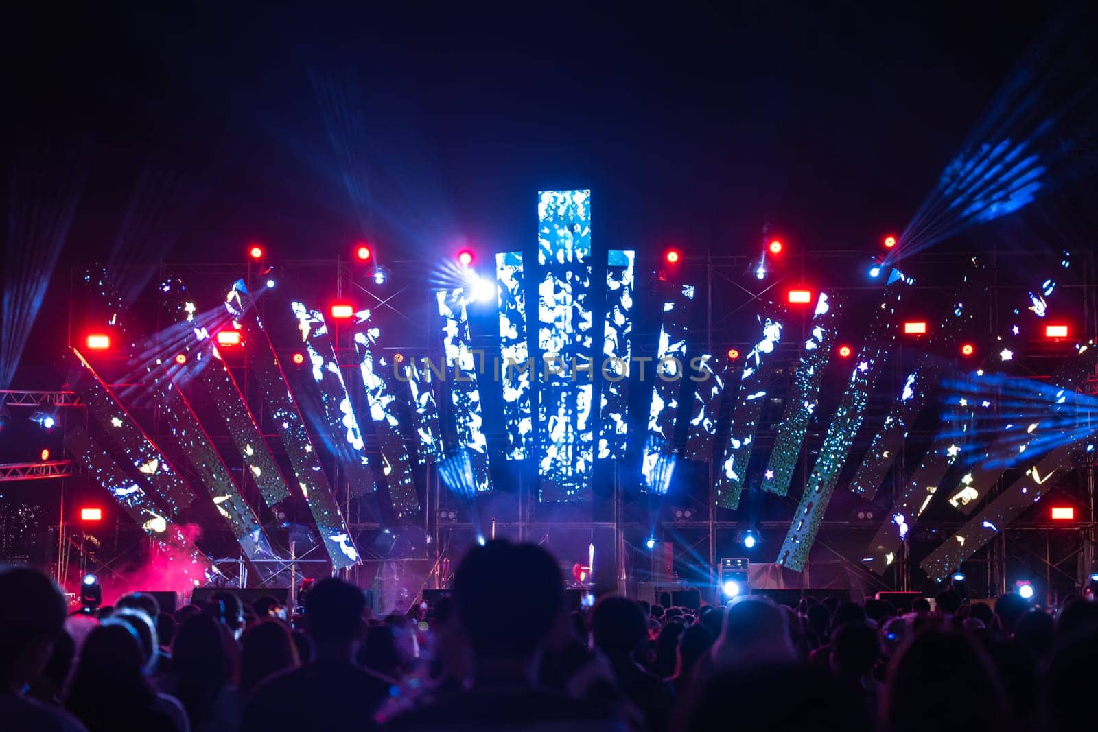 In midst of night a concert festival main event draws an enthusiastic unrecognizable crowd all cheering in front of brilliantly illuminated stage. lens flare adds to fun during this music festival.