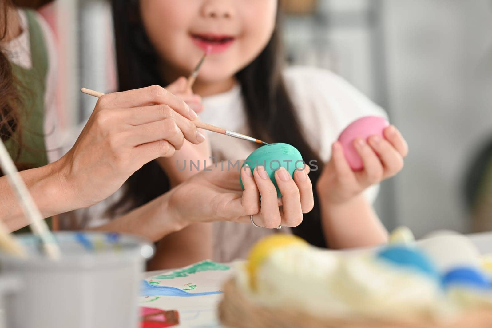 Smiling little girl painting eggs with mother at home, preparing for Easter by prathanchorruangsak