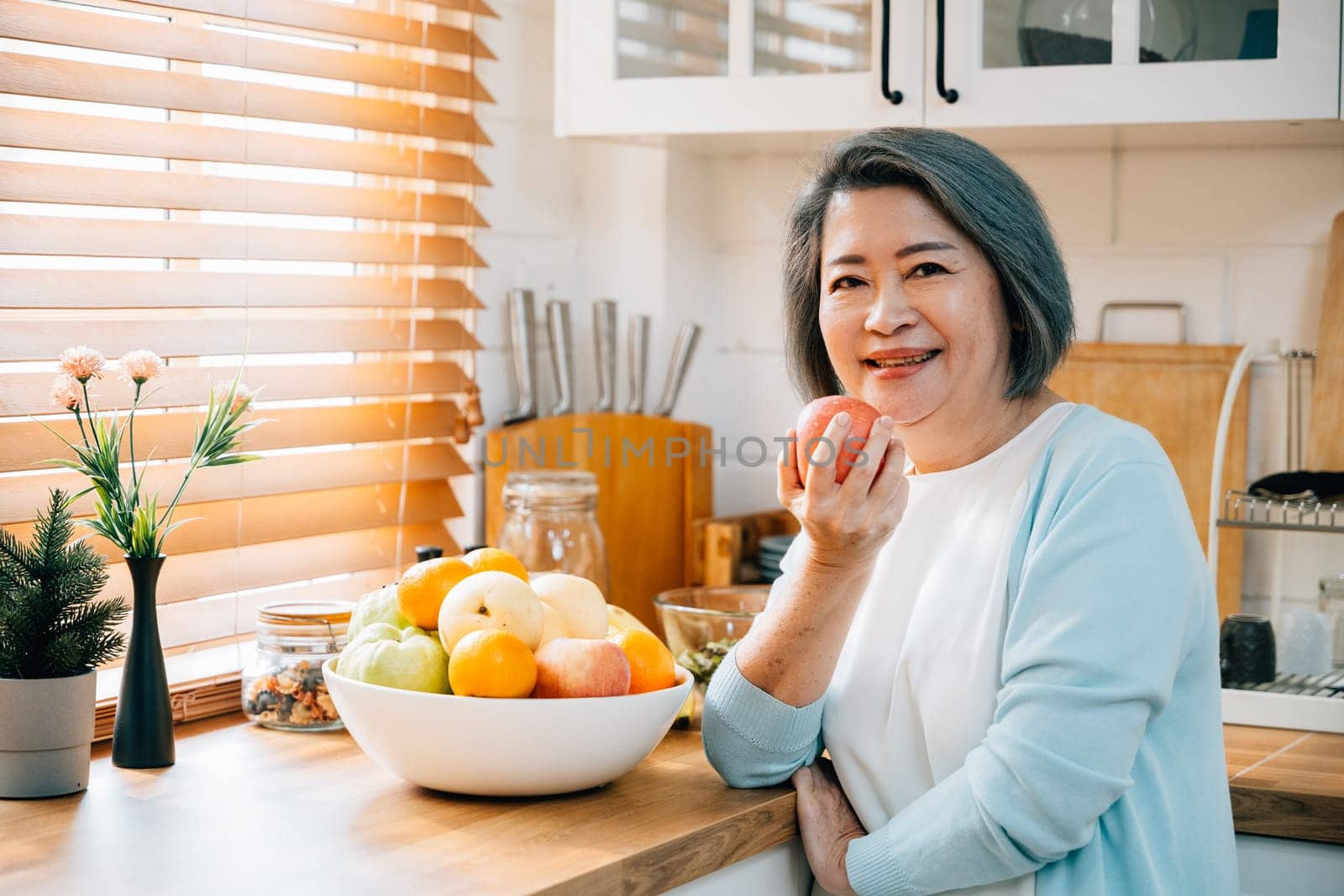 In the kitchen, a happy old woman, a grandmother, enjoys cooking and preparing healthy vegan food. She selects a fresh apple to eat, embodying the diet concept. A portrait of happiness and wellness. by Sorapop