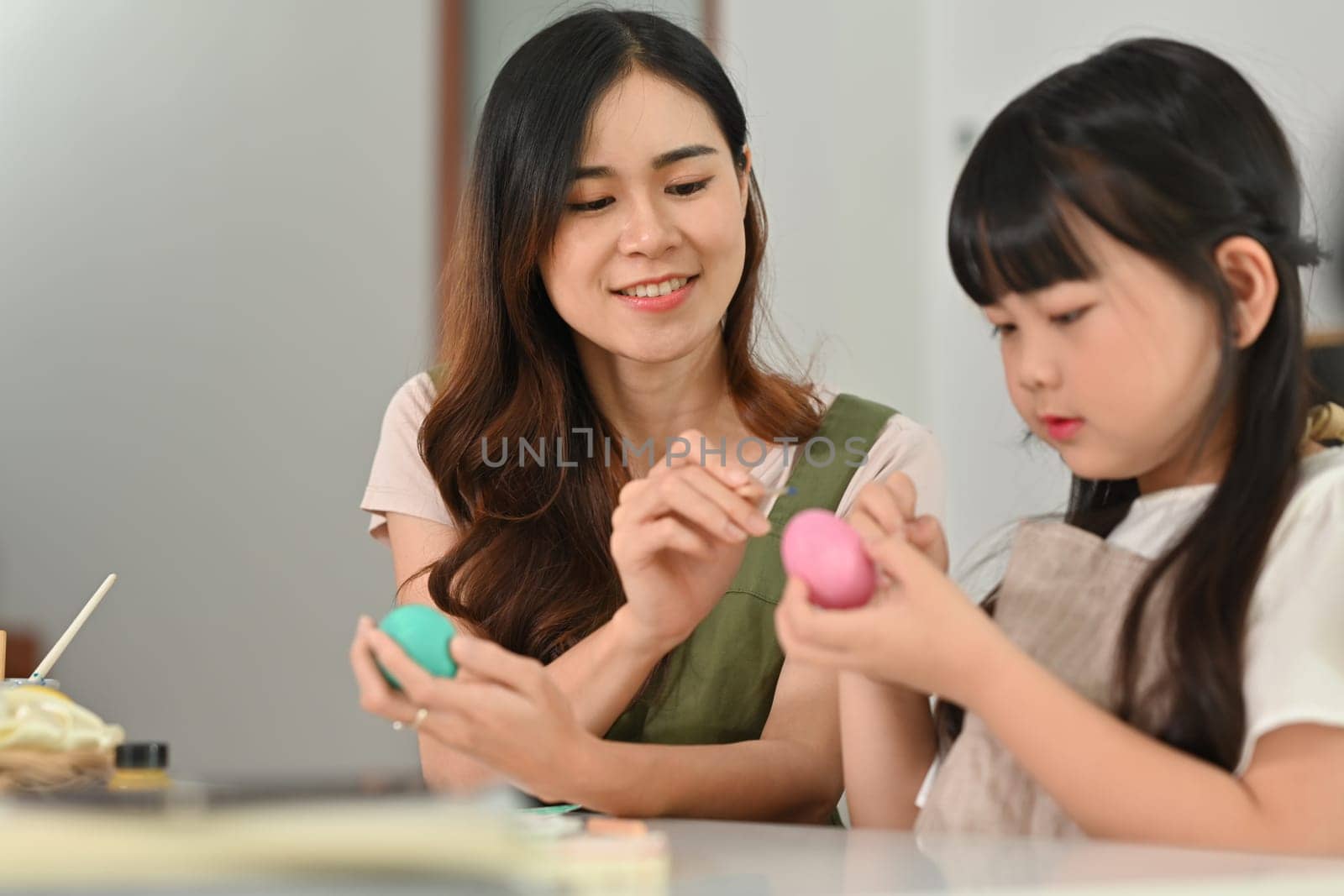 Smiling little girl painting Easter eggs with mother, getting ready for holiday at home.