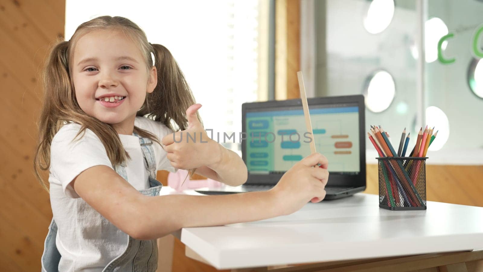 Smiling girl using laptop while look and show thumb up to camera. Cute child wearing headphone while working by using laptop writing code in STEM technology education. Online learning. Erudition.