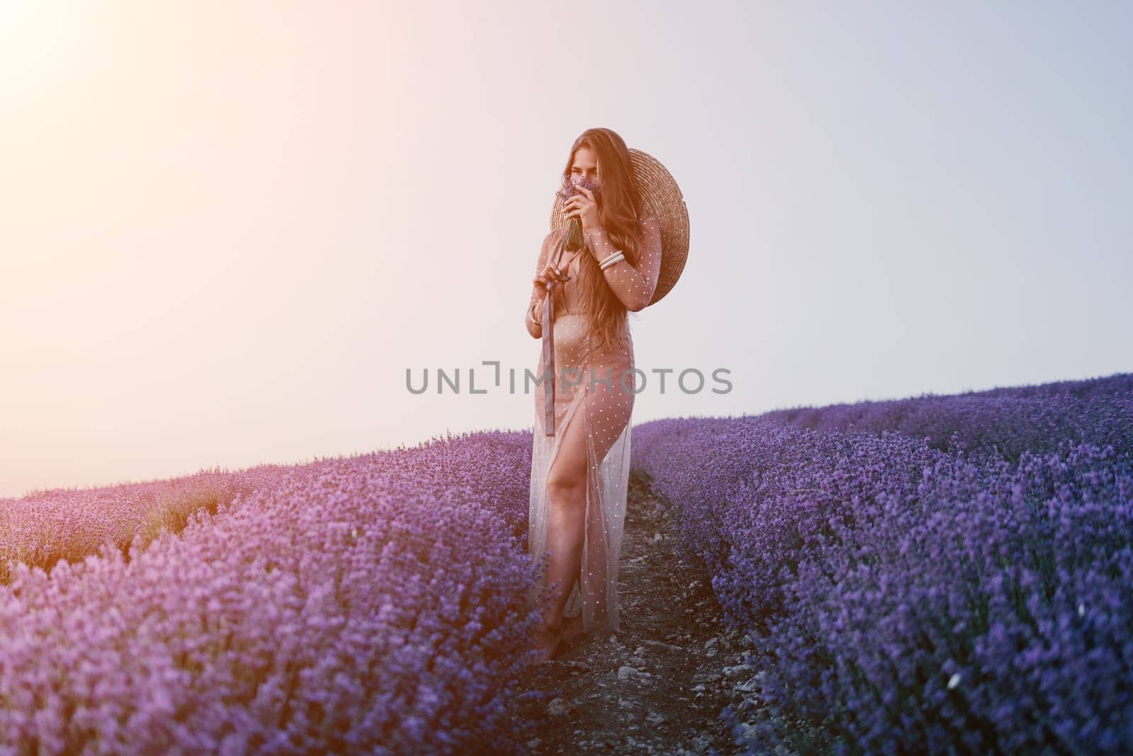 Close up portrait of young beautiful woman in a white dress and a hat is walking in the lavender field and smelling lavender bouquet.