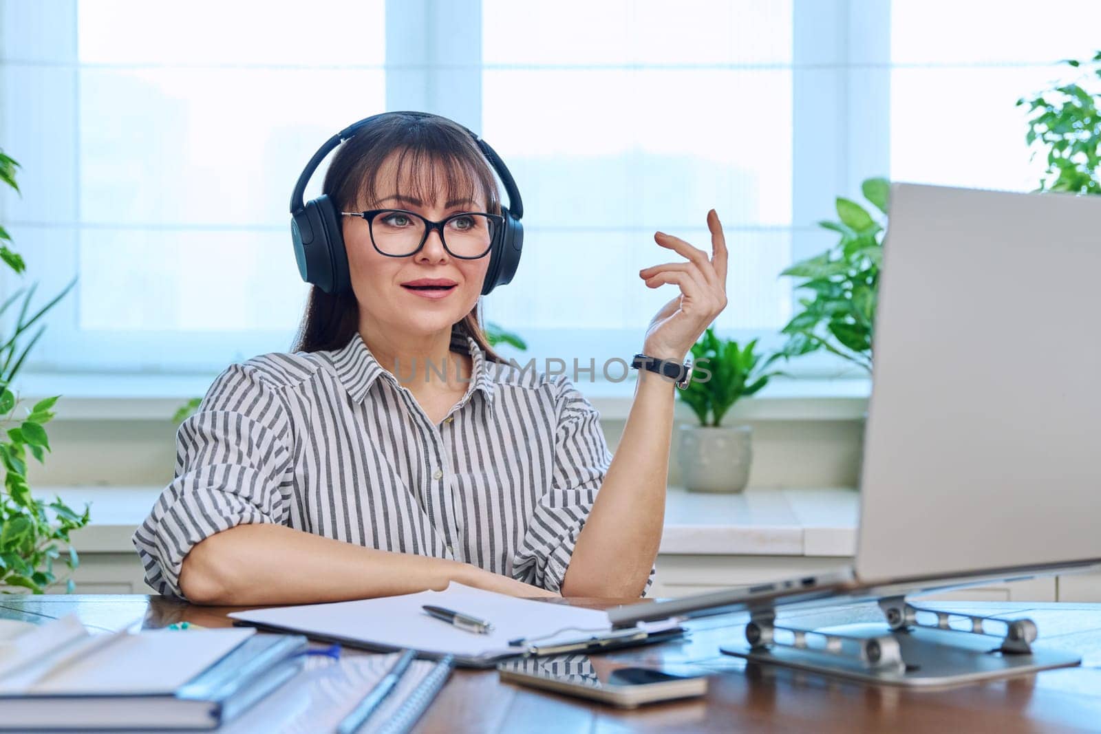 Middle-aged woman in headphones working at computer in home office, looking at web cam talking. Online meeting, remote work teaching financial legal advice interview blogging freelance, mental therapy