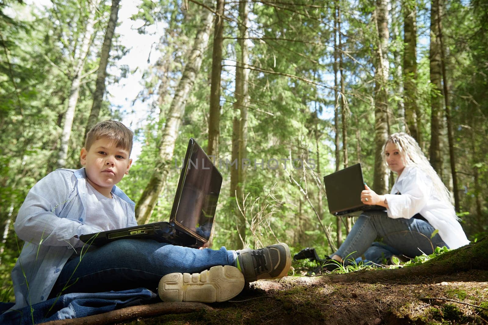 Mother and son with a laptops in the forest in summer. Fat young smart teenage boy and woman working with modern IT technologies in nature