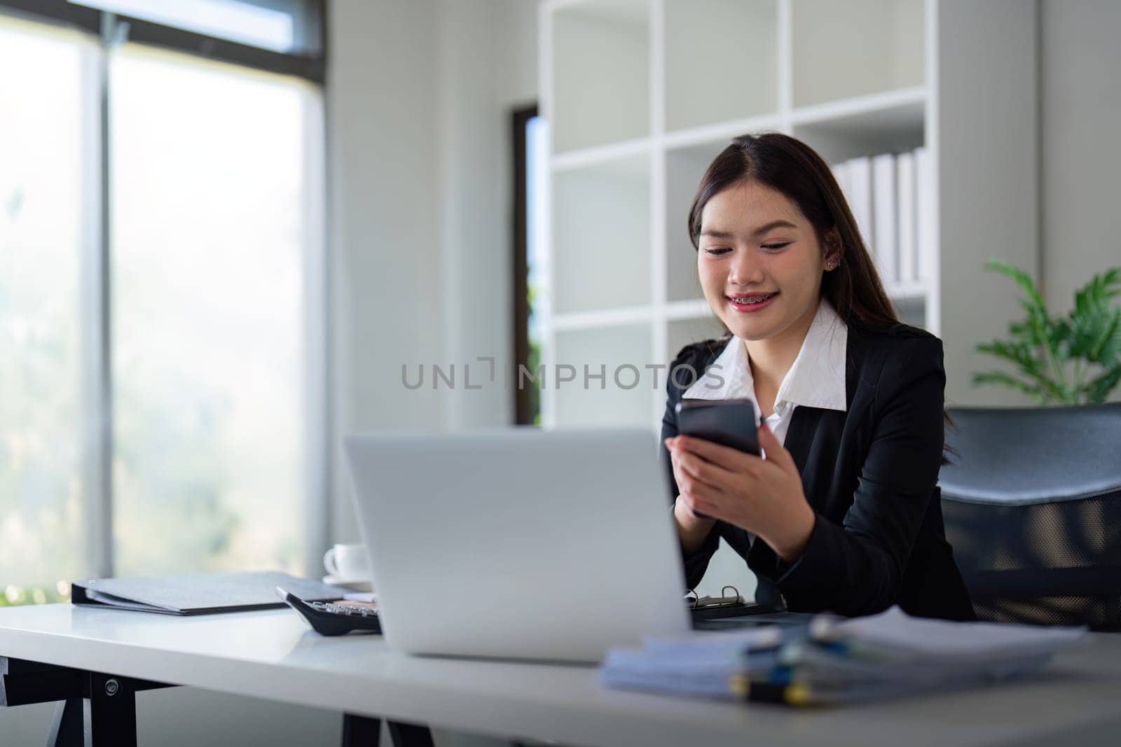 Beautiful young business woman using phone and laptop in office by nateemee