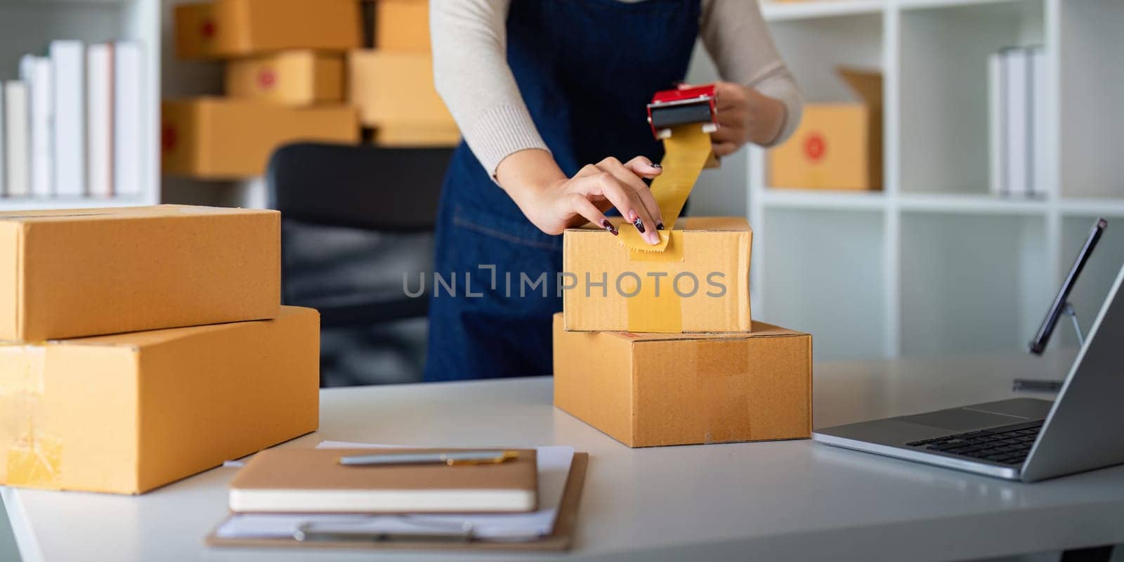 Woman use scotch tape to attach parcel box to prepare goods for the process of packaging, shipping, online sale internet marketing ecommerce concept startup business idea by nateemee