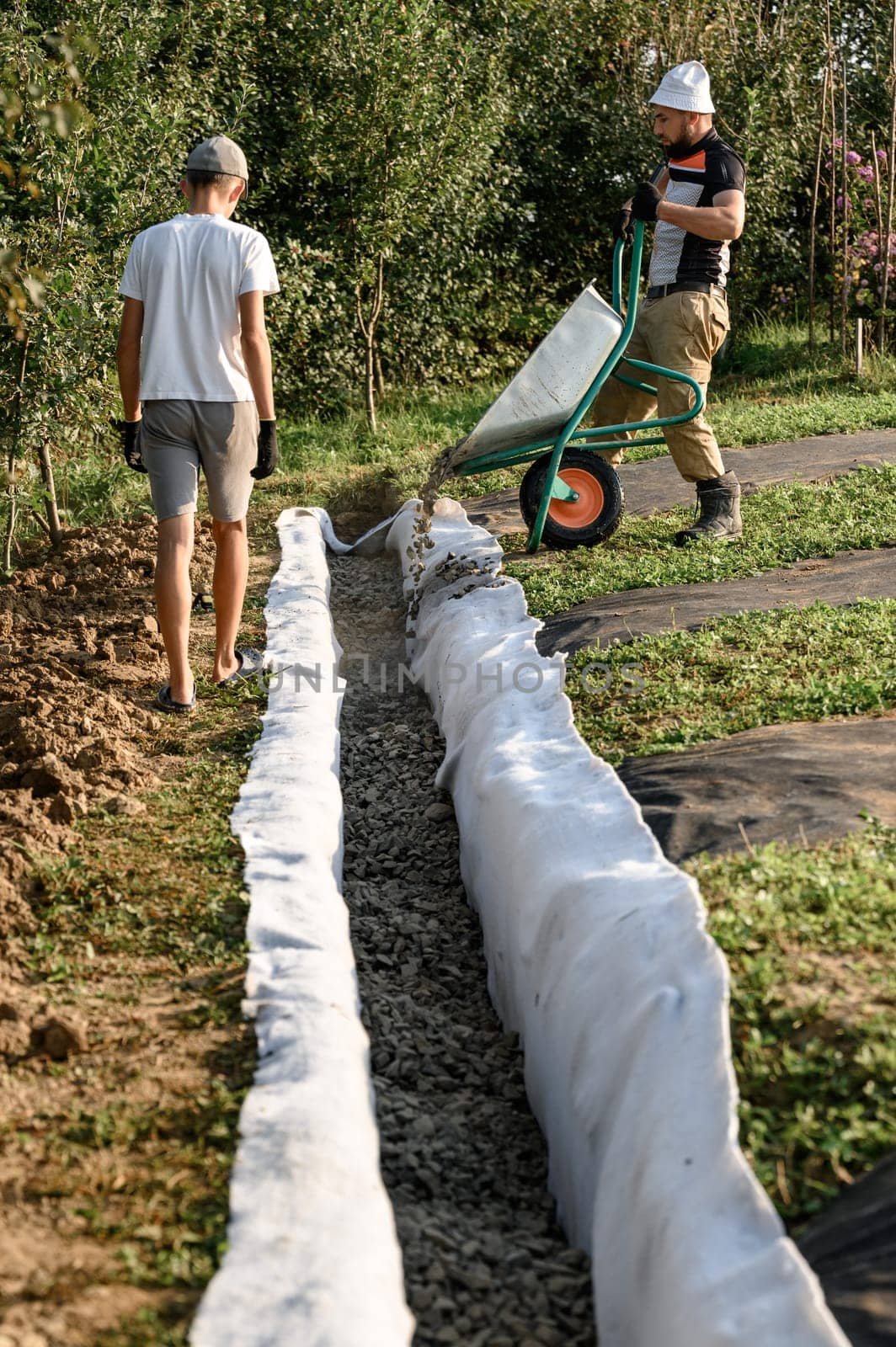 A man pours rubble crushed stone from a wheelbarrow into a trench. Drainage works for drainage of ground water around the field.