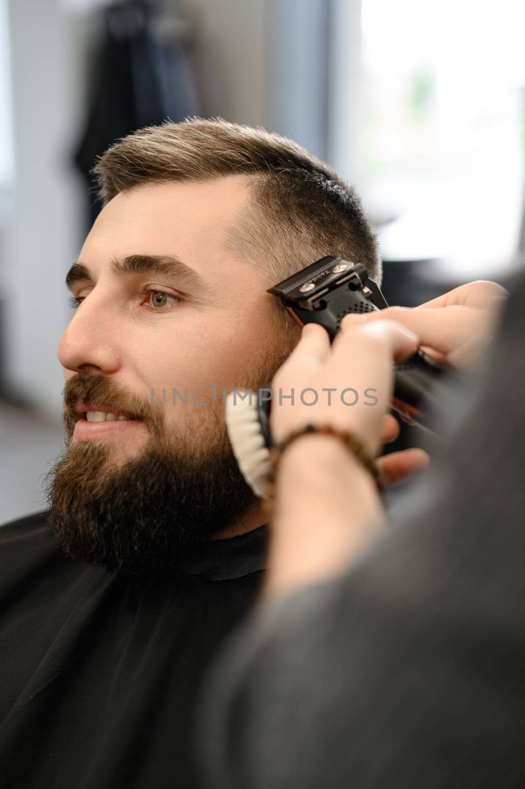 Barber shaves the contour of the oval line with a clipper on the clients head. A man with a beard gets a haircut in a barbershop chair.