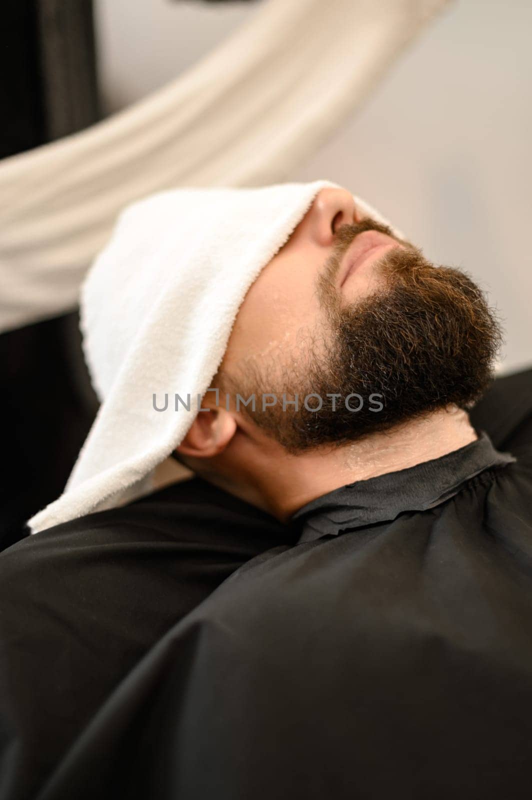A client is lying on a chair in a barbershop with his beard wrapped in a towel. A towel covers the man eyes. by Niko_Cingaryuk