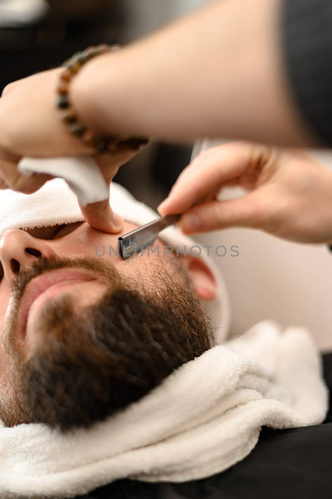 A barber shaves the cheek of a bearded customer with dangerous razor. Shaving the contour of the beard for the correct shape. by Niko_Cingaryuk