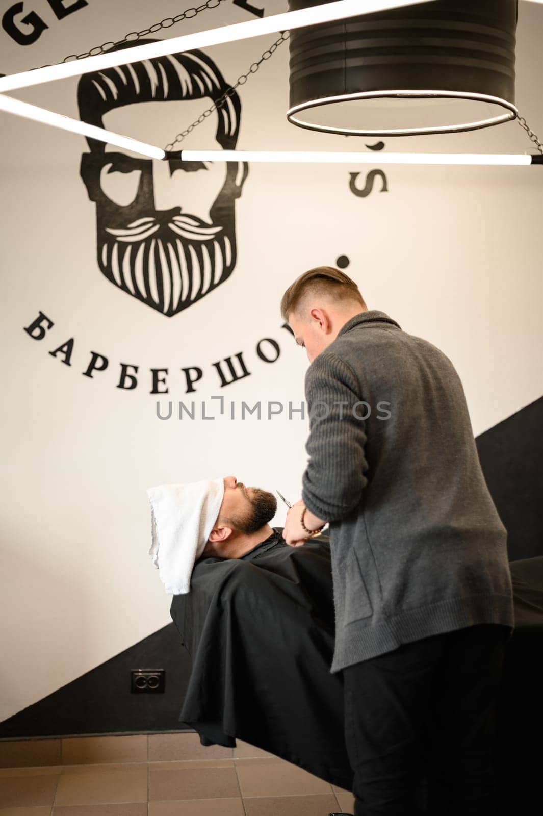 A barber stylist trims the beard of a Caucasian man, whose face is covered with a towel, with scissors