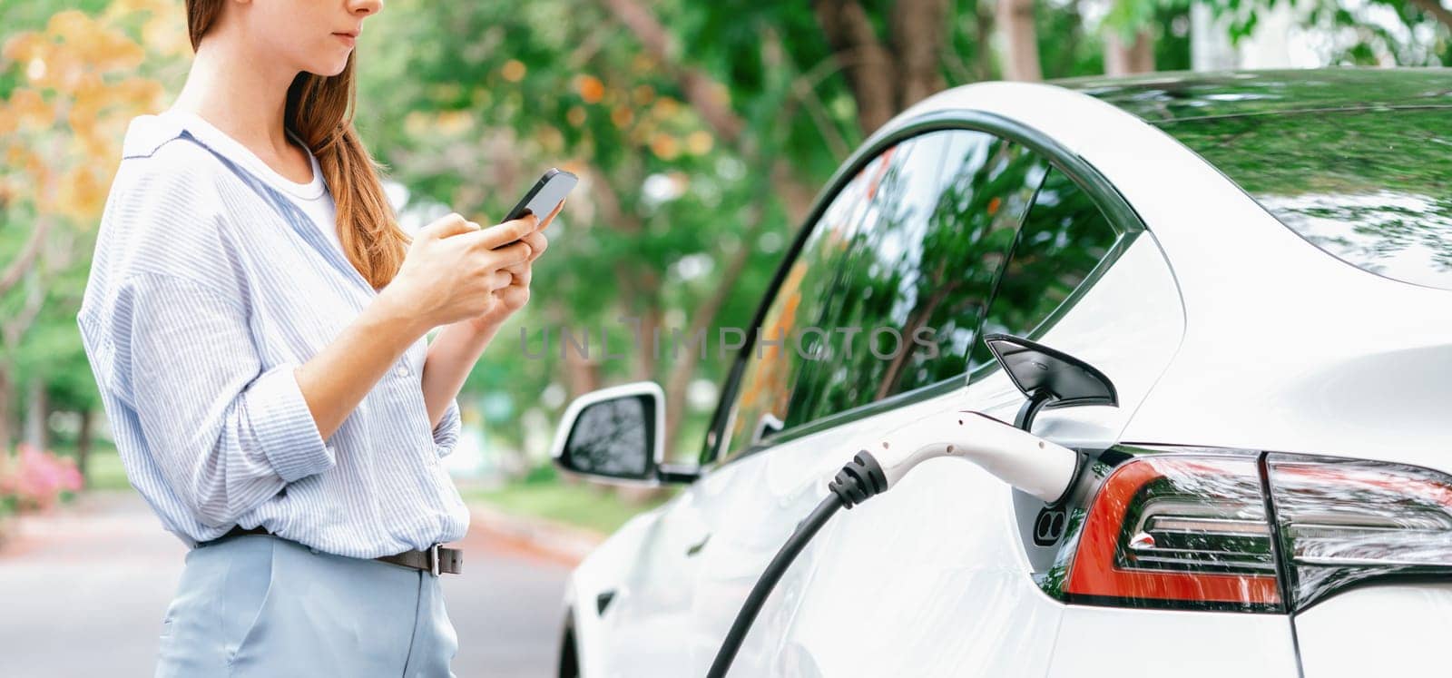 Panorama banner woman use smartphone online banking application to pay for EV car battery charging from EV charging station during autumn vacation holiday trip at national park or forest. Exalt
