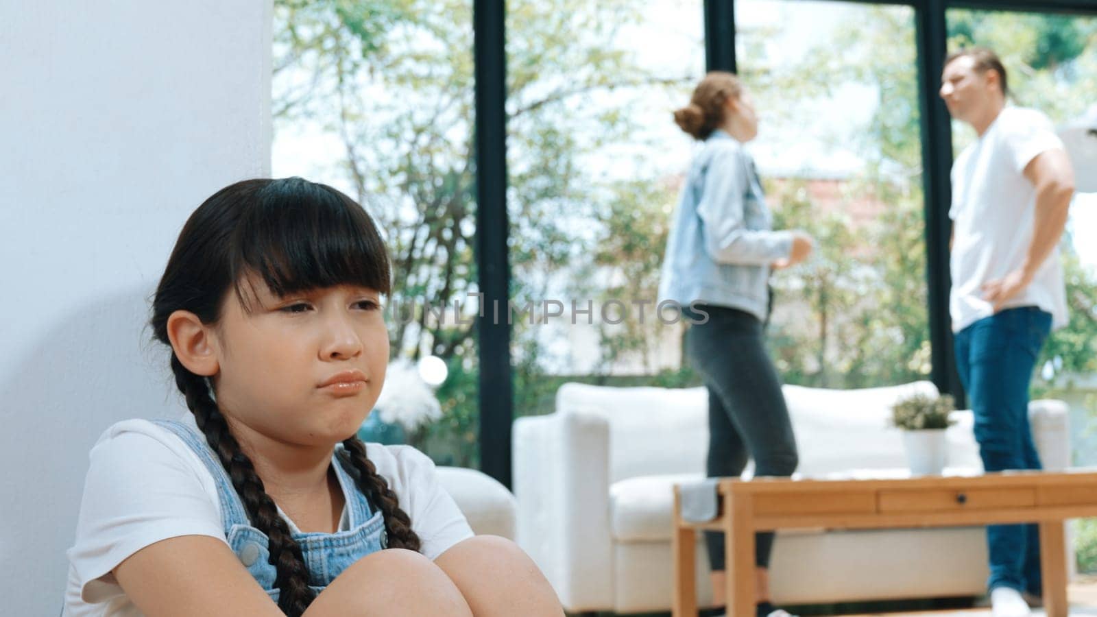 Stressed and unhappy young girl huddle in corner, cover her ears blocking sound of her parent arguing in background. Domestic violence at home and traumatic childhood develop to depression. Synchronos