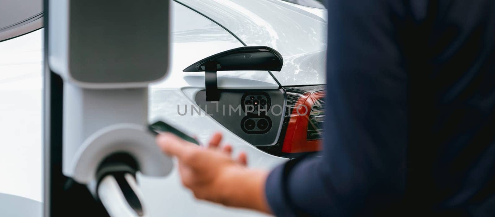 Man using smartphone online banking application to pay for electric car battery charging from EV charging station during vacation road trip at national park or summer forest. Panorama Exalt