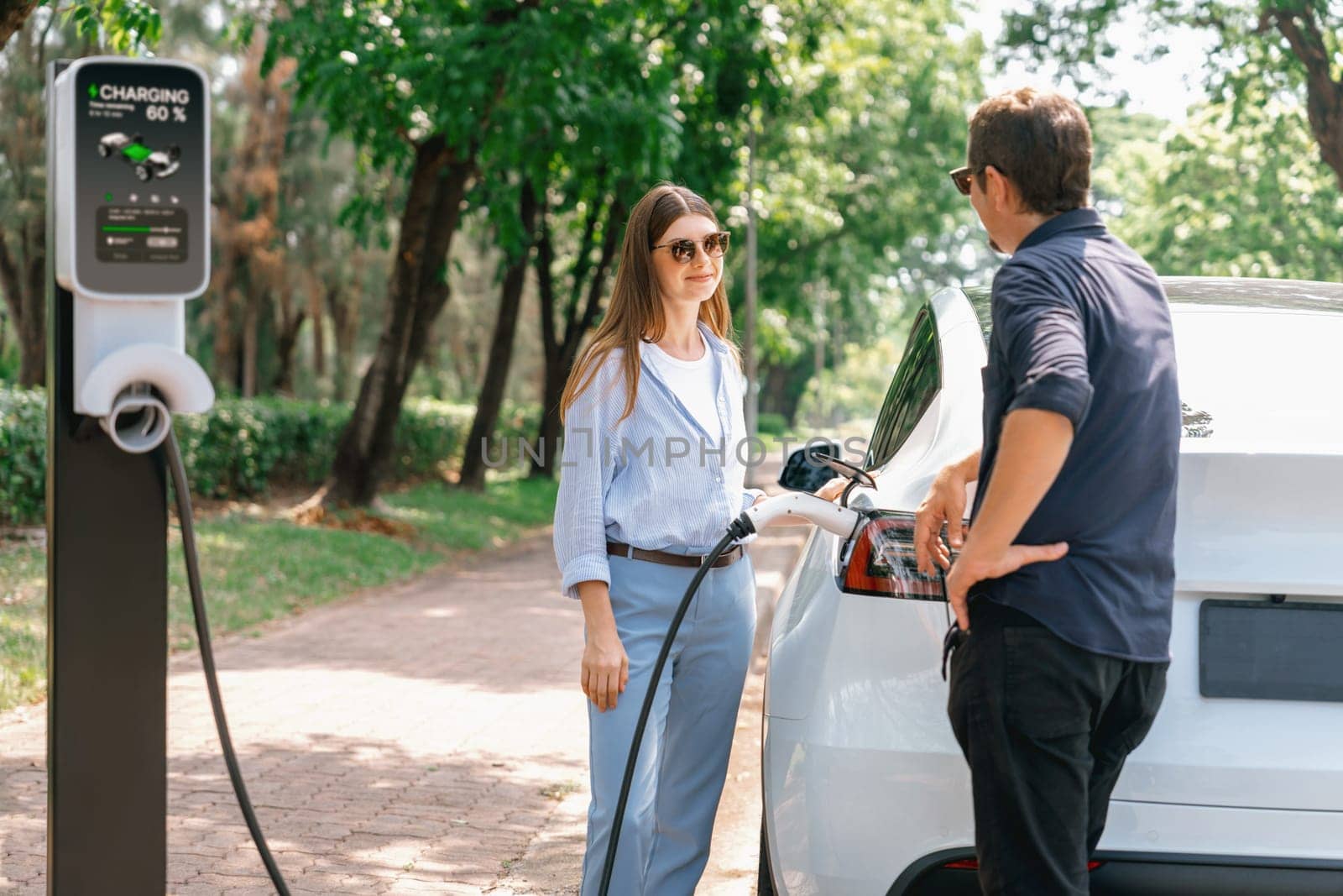 Lovely young couple wearing sun glasses recharging battery for electric car during road trip travel EV car in natural forest or national park. Eco friendly travel during vacation and holiday. Exalt