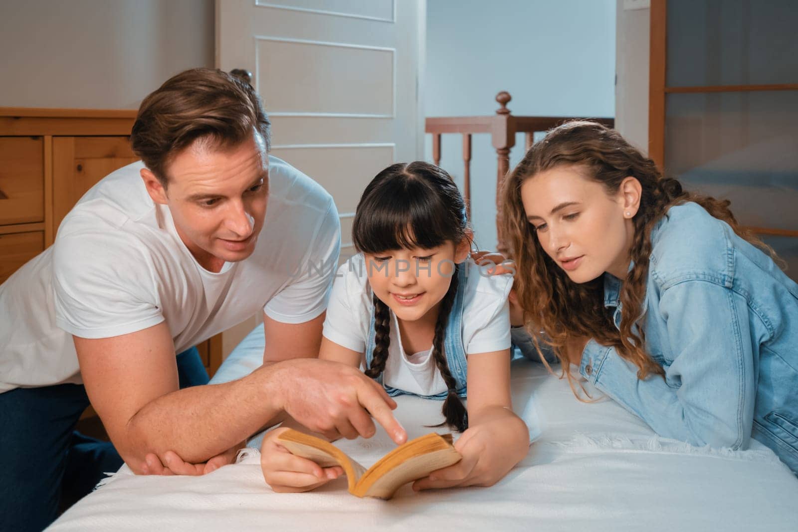 Bedtime story with childhood storytelling, mom and dad reading a fantasy book together to their little young girl in cozy and comfortable bedroom. Modern family happy time. Synchronos