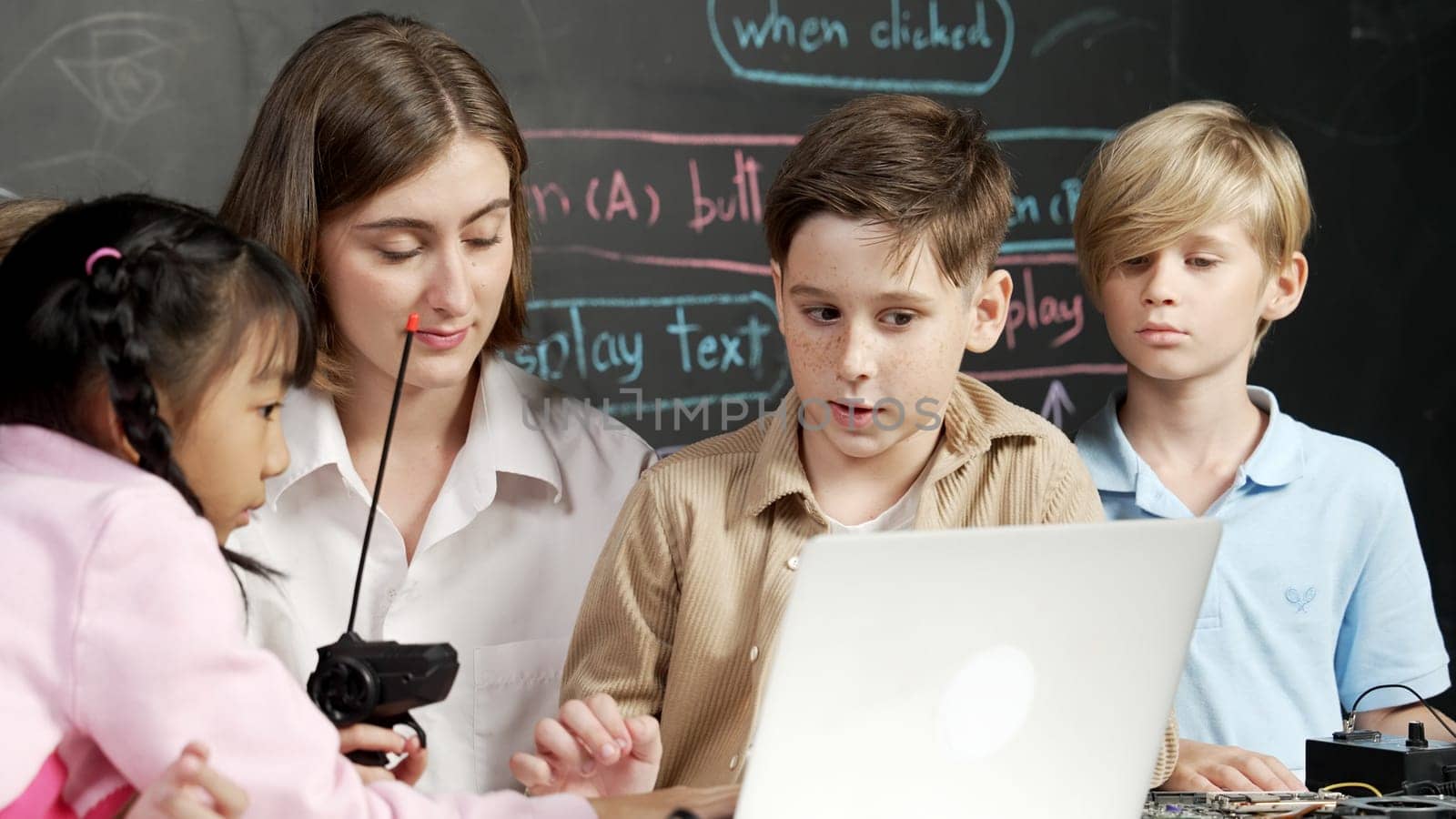 Caucasian boy using laptop programing engineering code and writing program while young caucasian teacher holding controller in STEM technology classroom at blackboard written with prompt. Erudition.