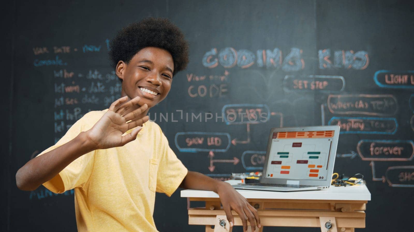 Smart african boy use laptop to code program and turn around to wave hand. Cute highschool stident coding engineering prompt or program system at blackboard in STEM technology classroom. Edification