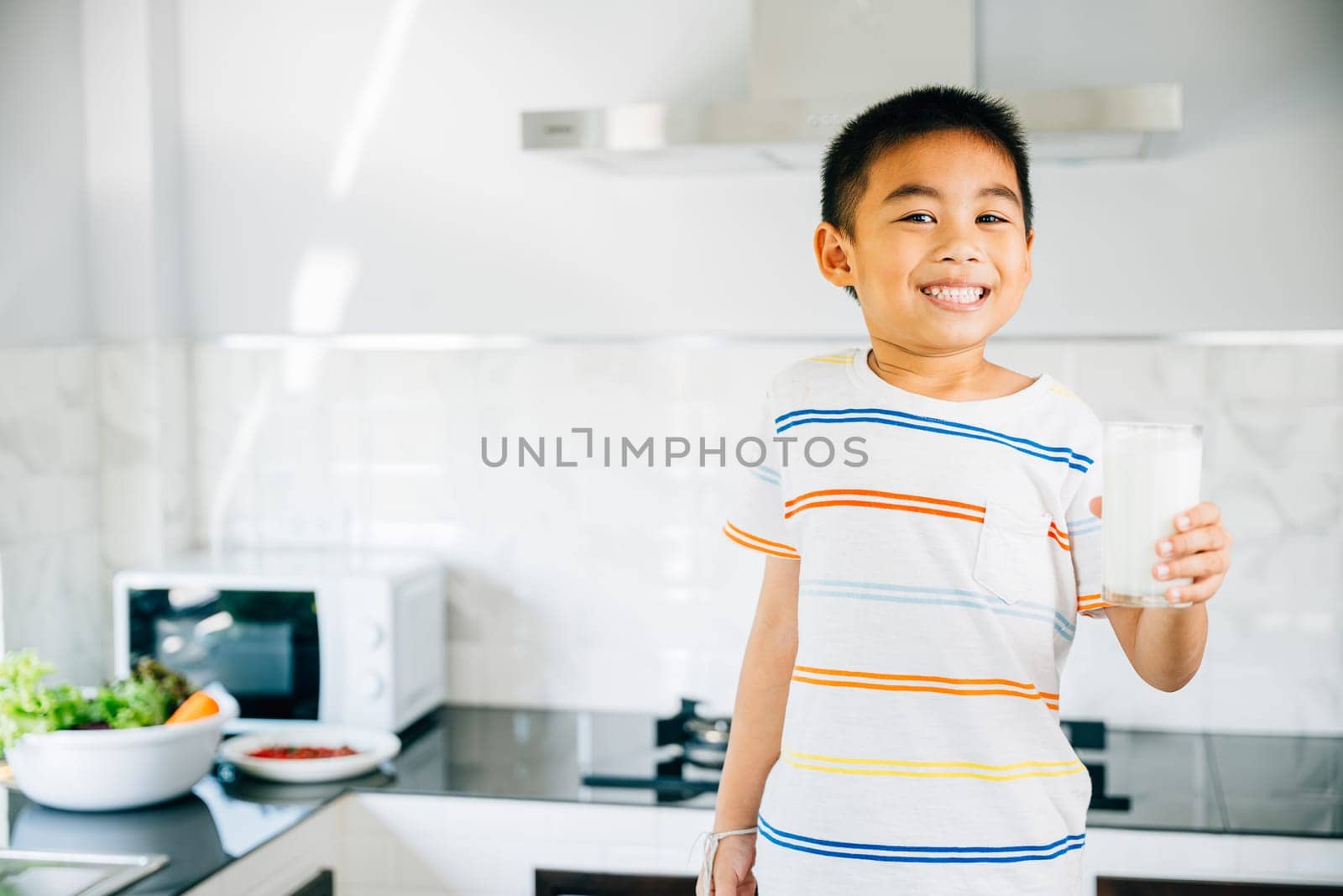 Portrait of adorable Asian preschooler, boy holds milk in kitchen. Smiling son enjoys drink, radiating joy. Happy child savoring calcium-rich liquid, feeling cheerful at home give me. by Sorapop