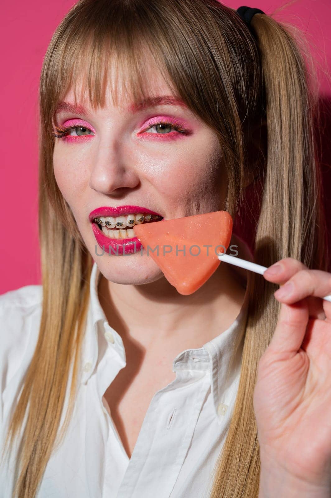 Portrait of a young woman with braces and bright makeup eating a lollipop on a pink background. Vertical photo. by mrwed54