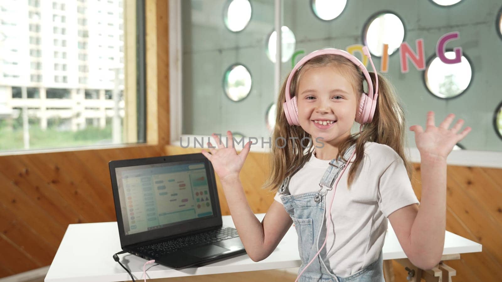 Smiling girl looking while waving hand at camera with laptop placed on table. Child wearing headphone smiling while laptop screen show system programing or coding program in STEM class. Erudition.