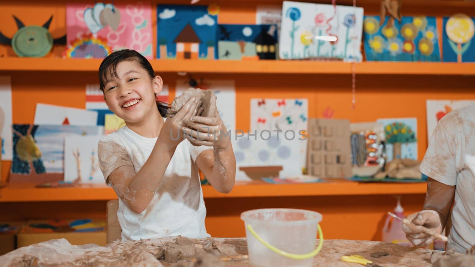 Asian highschool student modeling clay on table with mud and bottle of water at pottery workshop. Group of happy diverse children working or making cup of clay. Creative activity concept. Edification.