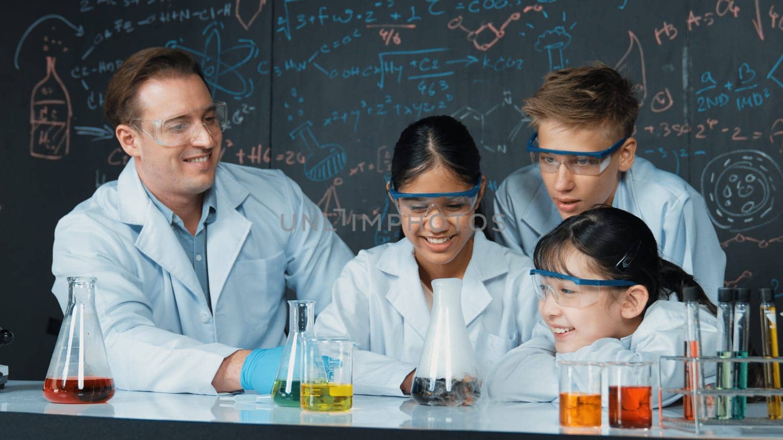 Girl looking at beaker while doing experiment at blackboard with chemistry theory in STEM class. teacher and group of highschool student with mixed races doing science activity together. Edification.