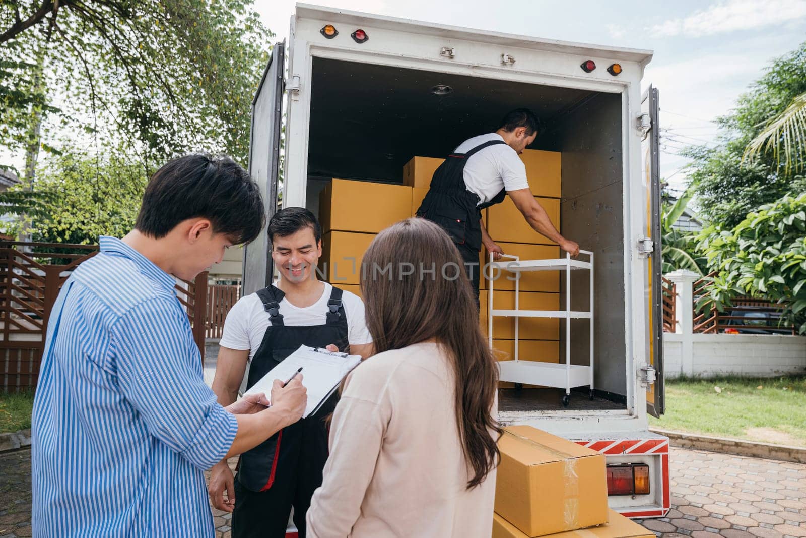 Professional movers assist a satisfied couple in signing the delivery checklist post-furniture handling. Uniformed employees prioritize customer happiness and satisfaction. Moving Day Concept
