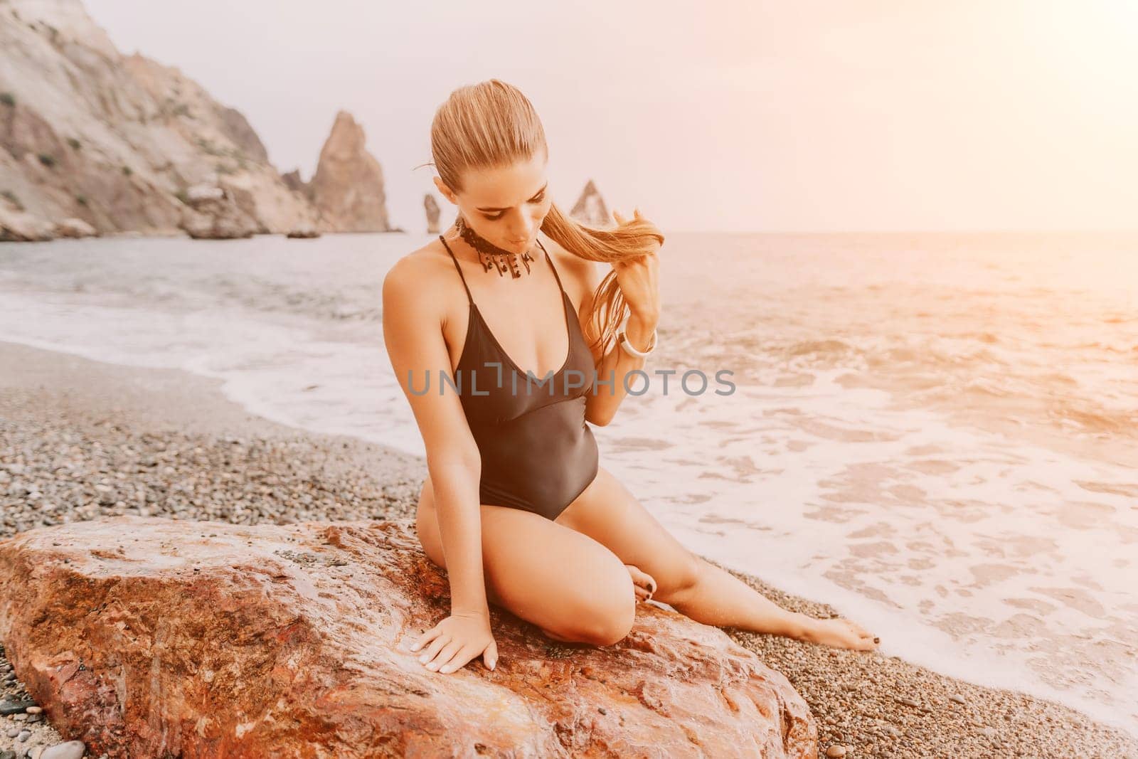 Woman travel portrait. Happy woman with long hair looking at camera and smiling. Close up portrait cute woman in black bikini posing on a red volcanic rock on the sea beach by panophotograph