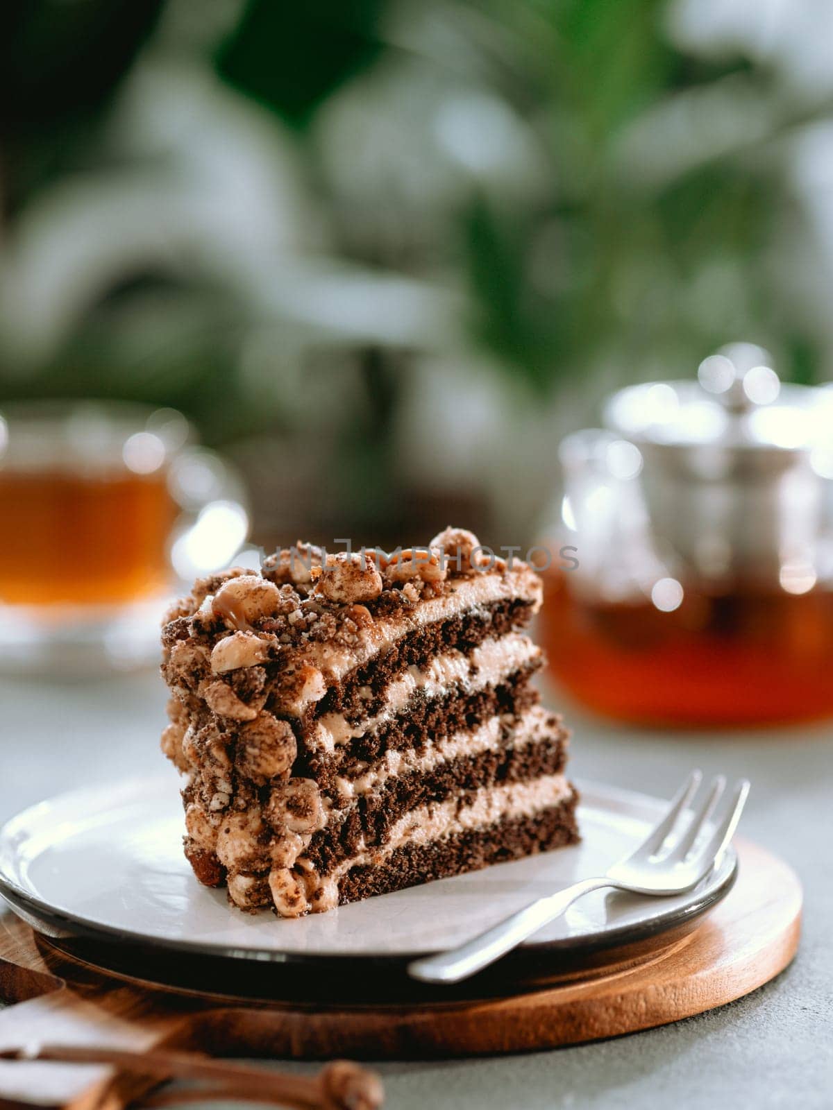 Perfect aesthetic layered cake with caramel and nuts on plate over gray table. Piece of chocolate almond biscuit with caramel frosting near tea cup and infuser teapot. Piece of sponge cake in cafe