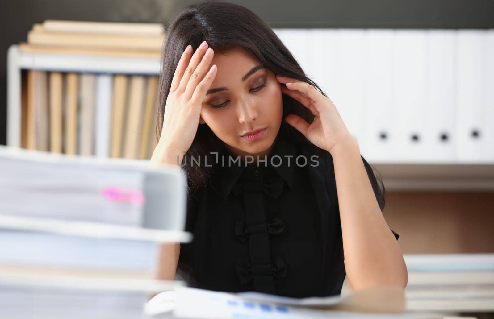 Tired and exhausted chinese woman looks at documents propping up her head with her hands. Huge pile of document folders, headache and depression concept