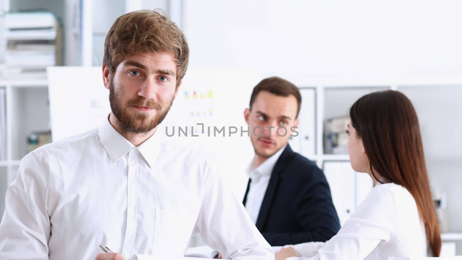 Handsome smiling bearded man in office look in camera with colleagues in background. White collar worker modern lifestyle graduate college study report idea coach train visit make note concept