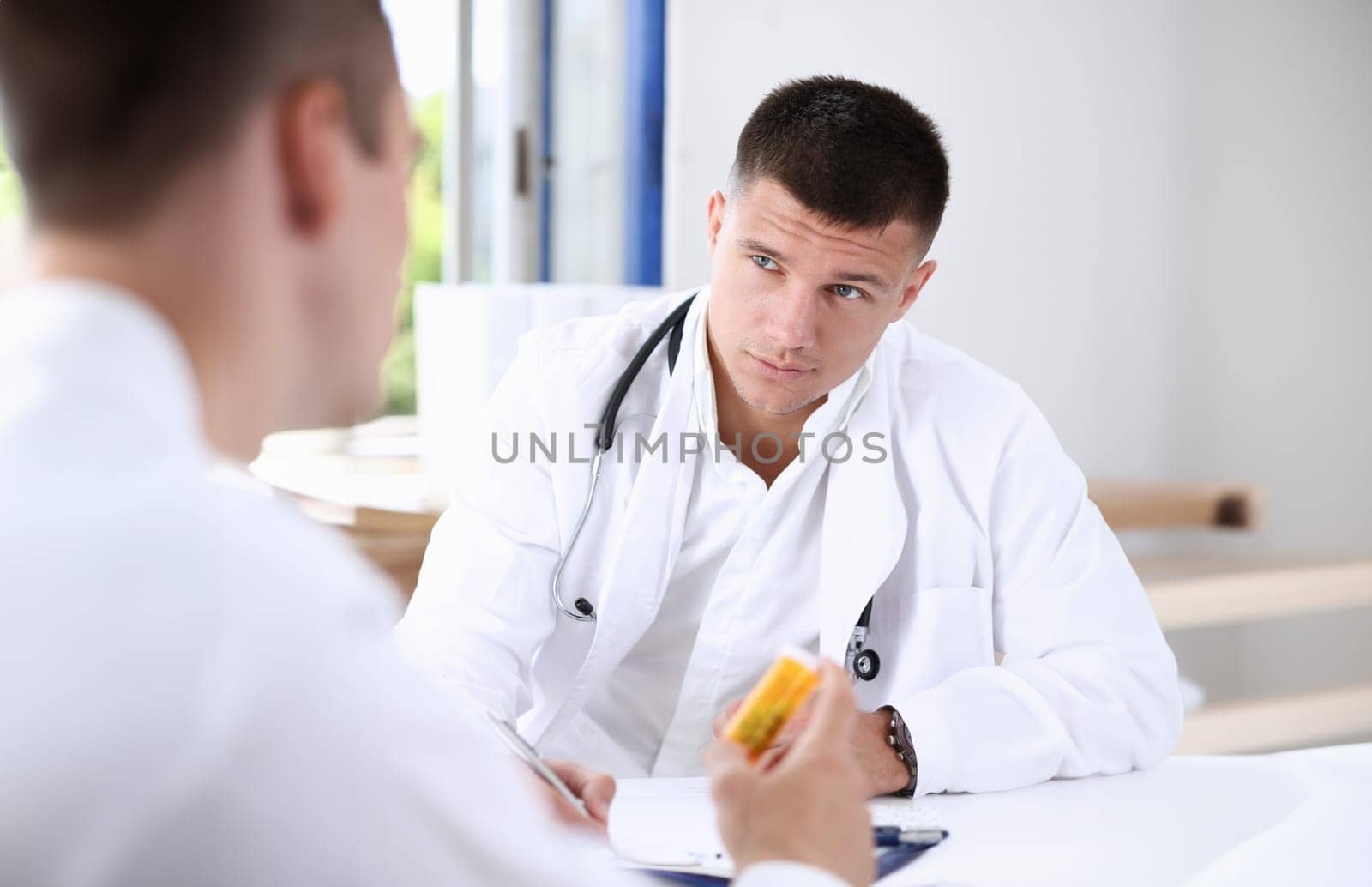 Male medicine doctor write prescription at worktable while patient hand hold jar of pills. Panacea and life save prescribing treatment legal drug store concept empty form ready to be used