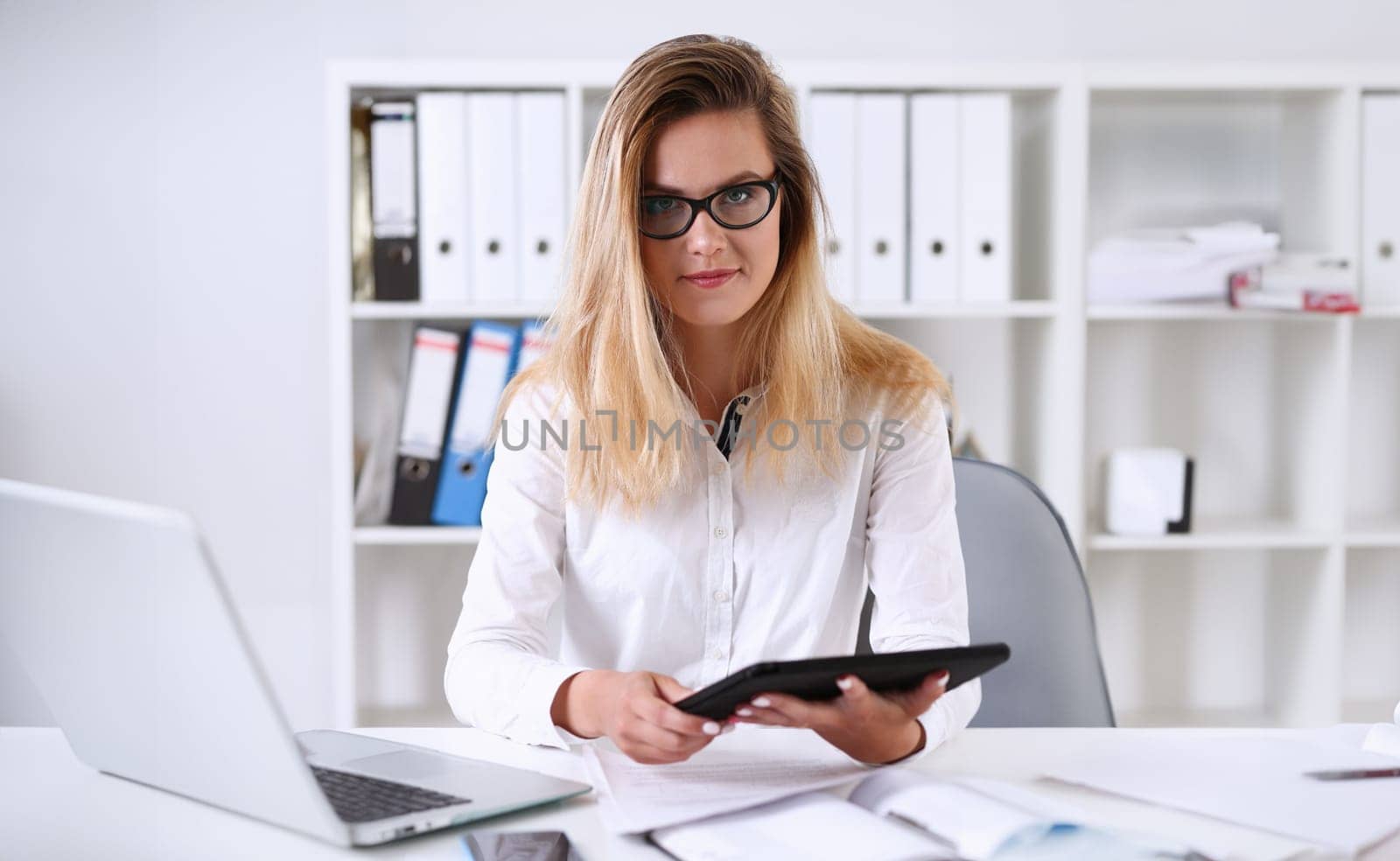 Beautiful businesswoman wearing glasses portrait at the office holding a tablet in hand sitting at the table smiling and looking at the camera teacher expresses success checking the test papers.